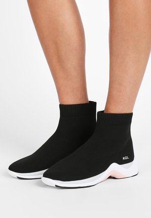 Baskets noires style chaussette avec semelles blanches et roses portées sur jambes nues, avec le logo « KGL » près du talon.