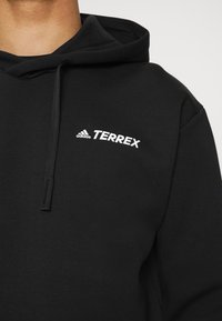 Sudadera negra de algodón, con un bolsillo canguro. Tiene una capucha con cordón y el logo blanco de "adidas Terrex" en el pecho.