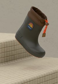 Botte imperméable noire et marron avec cordon orange et logo de montagne, positionnée au-dessus de marches en carrelage beige.