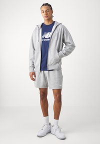 Vyras stovi dėvėdamas pilką užtrauktuku megztinį, pilkas šortus, tamsiai mėlyną New Balance marškinėlį, baltas kojines ir baltus Nike sportbačius ant balto fono.