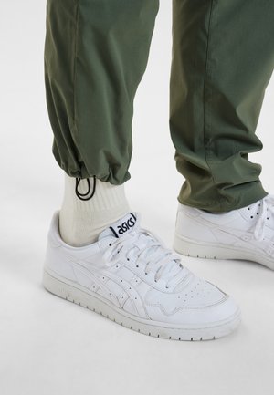 Zapatillas de cuero blancas con diseño perforado y acentos grises, combinadas con pantalones verdes ajustados con puños elásticos y calcetines beige de caña corta.