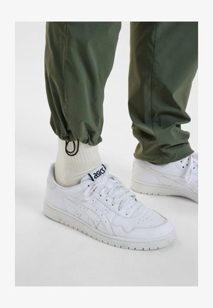 Witte leren sneakers met een geperforeerd ontwerp en grijze accenten, gecombineerd met groene taps toelopende broek met elastische boorden en beige crew-sokken.