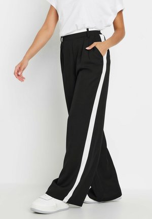 Persona che indossa pantaloni neri a gamba larga con strisce laterali bianche e scarpe da ginnastica bianche, in piedi con una mano in tasca.