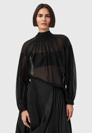 Donna con capelli corti scuri che indossa una blusa nera trasparente con collo alto sopra un top bandeau nero e una gonna plissettata nera, su uno sfondo chiaro.