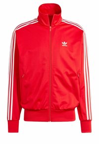 Punainen Adidas-takki, jossa korkea kaulus, vetoketju edessä, valkoiset sivuraidat, kaksi vetoketjullista taskua ja pieni logo rinnassa. Sileä pinnan rakenne.