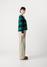 Gestreepte langhandsleeve top in groen en marineblauw, gecombineerd met lichtbeige wijde broek. Bruine instap-schoenen met veters. Gladde texturen.