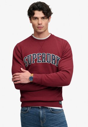 Mann mit lockigem dunklem Haar, der einen dunkelroten Superdry-Sweatshirt und blaue Jeans trägt, mit verschränkten Armen steht und eine beige Lederuhr trägt.