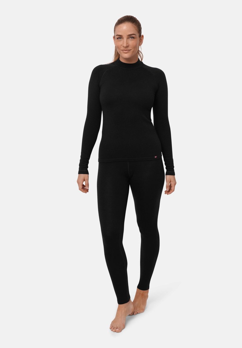 Frau steht barfuß in einem enganliegenden schwarzen Langarmshirt und passenden Leggings vor einem einfachen weißen Hintergrund.