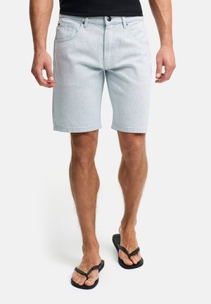Mann iført lyseblå denimshorts og svarte flip-flops, stående mot en enkel hvit bakgrunn.