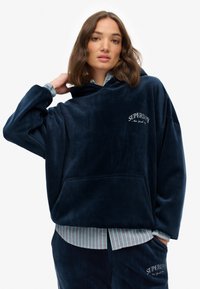 Sudadera con capucha en azul marino, con un gran bolsillo frontal, de corte holgado y logotipo bordado de 'Superdry'. Lleva una camiseta de rayas debajo.
