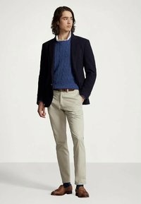 Άνδρας που στέκεται φορώντας μπλε σακάκι, μπλε πλεκτό πουλόβερ, μπεζ παντελόνι, καφέ ζώνη και καφέ φλατ παπούτσια σε λευκό φόντο.