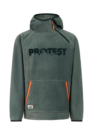 Jonge persoon die een grijze gebreide muts draagt, een groene hoodie, feloranje broek en zwarte laarzen, staand met handen in de zakken.