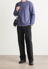 Lila sweatshirt med rund halsringning, ribbade ärmslut och nederkant. Kombinerad med svarta byxor och svart axelremsväska, står på ett ljust golv.