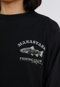 Black cotton t-shirt featuring a graphic of a fish and the text "MANASTASH FISHING CLUB" in white. Short sleeves and crew neck design.