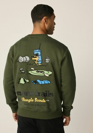 Olivgrüner Pullover mit einem Cartoon-Design auf dem Rücken, das Camping-Elemente zeigt, darunter einen Hund, Zelte und Kajaks, mit Text darunter.