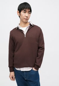 Brun genser med glidelås i teksturert stoff, med en hvit undertrøye og en lys stripete krage. Brukt sammen med blå denimjeans.
