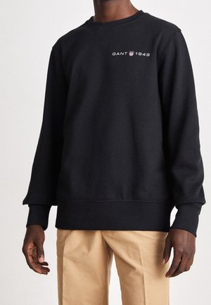 Man wearing a black sweatshirt with "GANT 1949" logo on the chest and beige pants, standing against a plain light background.