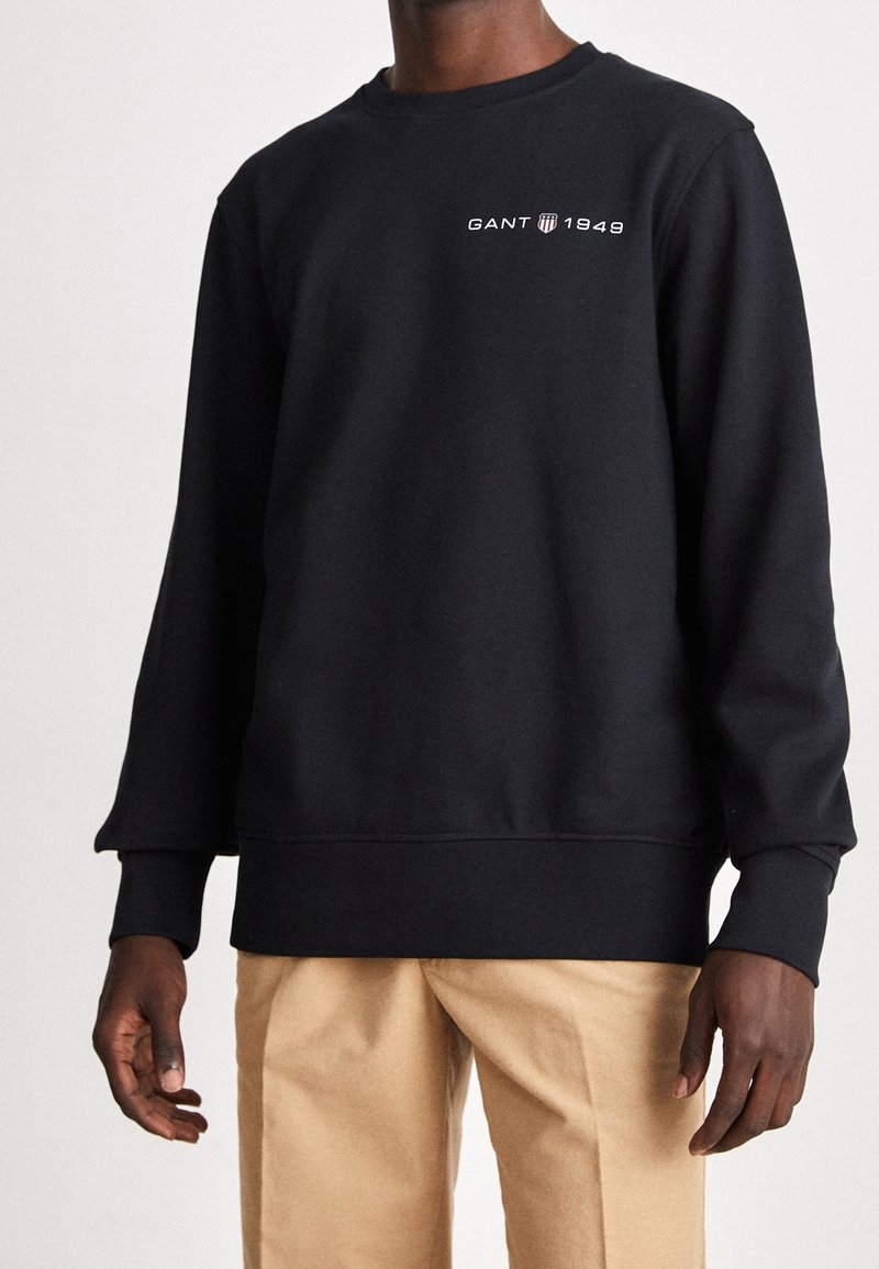 Homme portant un sweat-shirt noir avec le logo "GANT 1949" sur la poitrine et un pantalon beige, debout devant un fond clair uni.