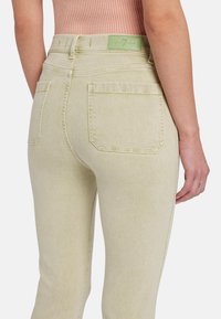 Lichtgele denim jeans hebben een hoge tailleband, twee achterzakken en subtiele stiksels. Een groene merklabel is zichtbaar bij de taille.