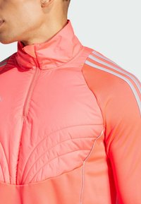 Roze sportpullover met een opstaande kraag, voorzien van gestepte details op de borst en lichtgrijze strepen op de schouders.