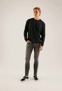 Giovane in piedi con le mani in tasca, indossando una maglietta nera a maniche lunghe, jeans skinny grigi strappati e sneakers nere con strisce bianche.