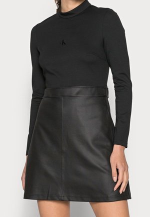 Donna che indossa una maglia nera a manica lunga con collo alto finto e piccolo logo "ck" e una gonna nera in ecopelle a vita alta.