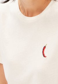 Krämfärgad bomulls-t-shirt med en röd och grön broderad chilipeppar-accent på vänster bröstområde. Korta ärmar och rundad halsringning.