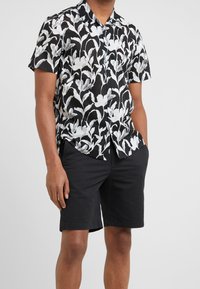 Man i en svart och vit blommig kortärmad skjorta med knappar, tillsammans med svarta knälånga shorts, stående med händerna i fickorna.