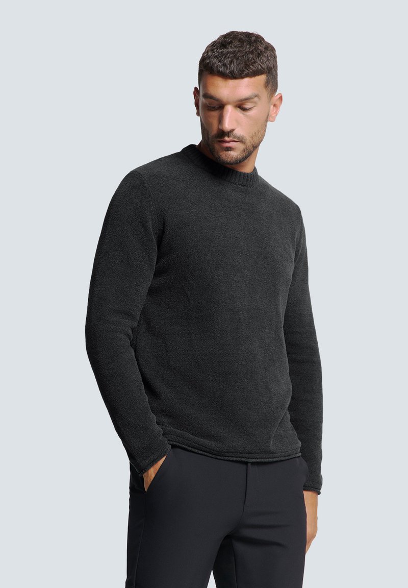 Schwarzer Rundhalsausschnitt-Pullover aus strukturiertem Stoff, mit langen Ärmeln und lockerer Passform. Getragen mit dunklen Hosen vor einem hellen Hintergrund.