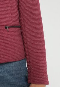 Vinröd texturerad jacka med ribbat tyg, med en dragkedja ficka fram och en figurnära design, ihop med en denimkjol.