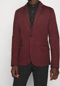 Bordowa marynarka wykonana z fakturowanej tkaniny, z pojedynczym zapięciem na guzik, klapami z wycięciem i dwoma przednimi kieszeniami, noszona na czarnej koszuli.