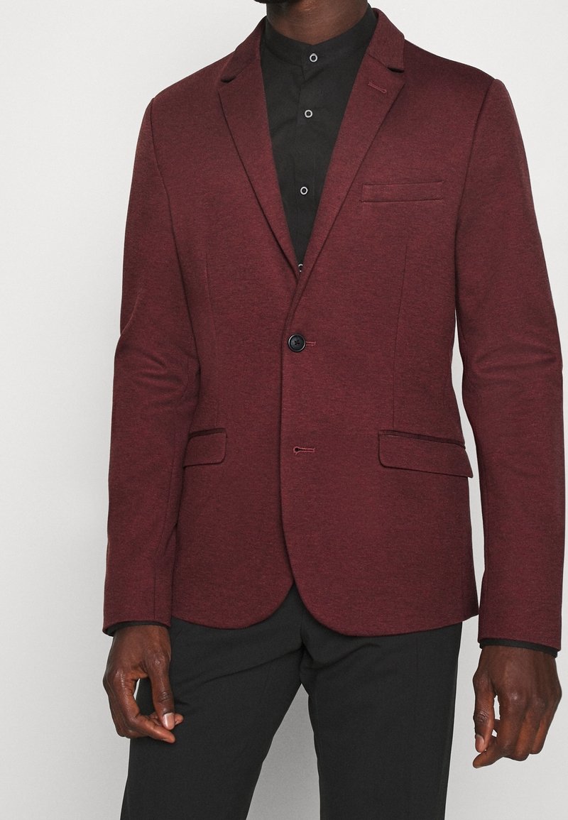 Bordowa marynarka wykonana z fakturowanej tkaniny, z pojedynczym zapięciem na guzik, klapami z wycięciem i dwoma przednimi kieszeniami, noszona na czarnej koszuli.