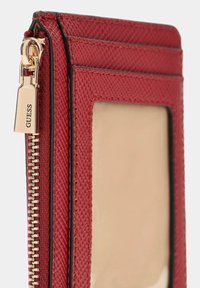 Red textured wallet with gold zipper pull engraved "GUESS," multiple card slots, and a transparent ID window on a white background.
