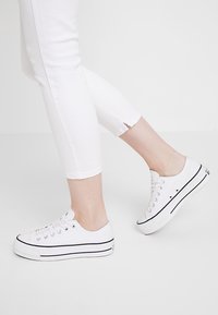 Scarpe da ginnastica in tela bianca con punta tonda, striscia nera lungo la suola e lacci bianchi. Indossate con pantaloni bianchi corti che lasciano scoperta la caviglia.