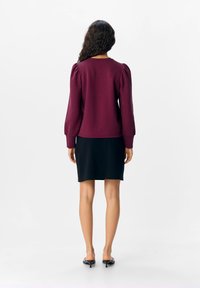 Burgunderfarbene Oberteil mit Puffärmeln, kombiniert mit einem schwarzen, knielangen Rock, der eine glatte Textur und eine taillierte Passform aufweist. Schwarze High Heel Sandalen runden das Outfit ab.