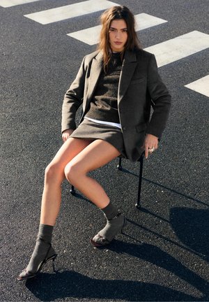 Donna con blazer marrone, maglione e gonna con calzini e tacchi, seduta su una sedia sull'asfalto vicino alle strisce pedonali.