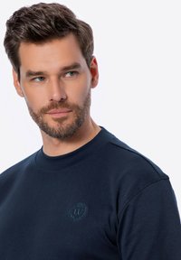 Námořnický modrý svetr vyrobený z měkké bavlny, s kulatým výstřihem a malým vyšívaným logem na hrudi. Hladká textura a uvolněný střih.
