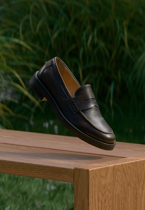 Zapato mocasín de cuero negro flotando sobre un banco de madera con plantas verdes desenfocadas al fondo.