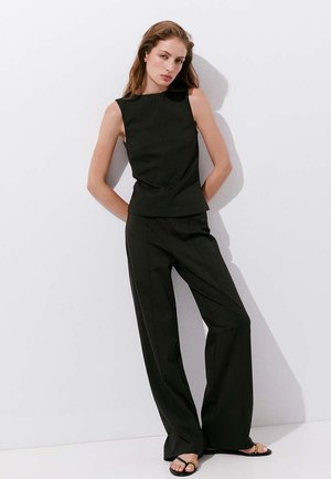 Femme aux longs cheveux portant un haut noir sans manches, un pantalon noir à jambes larges et des sandales noires, debout contre un mur blanc avec les mains dans le dos.