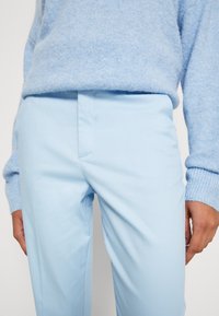 Světlé modré kalhoty na míru vyrobené z hladké látky, se zapuštěnými předními kapsami a štíhlým střihem, spárované s jemným svetrém v světlé modré barvě.