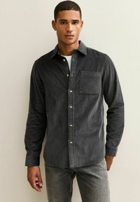 Camisa de corduroy gris oscuro con botones, un único bolsillo en el pecho, rayas verticales prominentes y botones dorados. Mangas largas y corte ajustado.