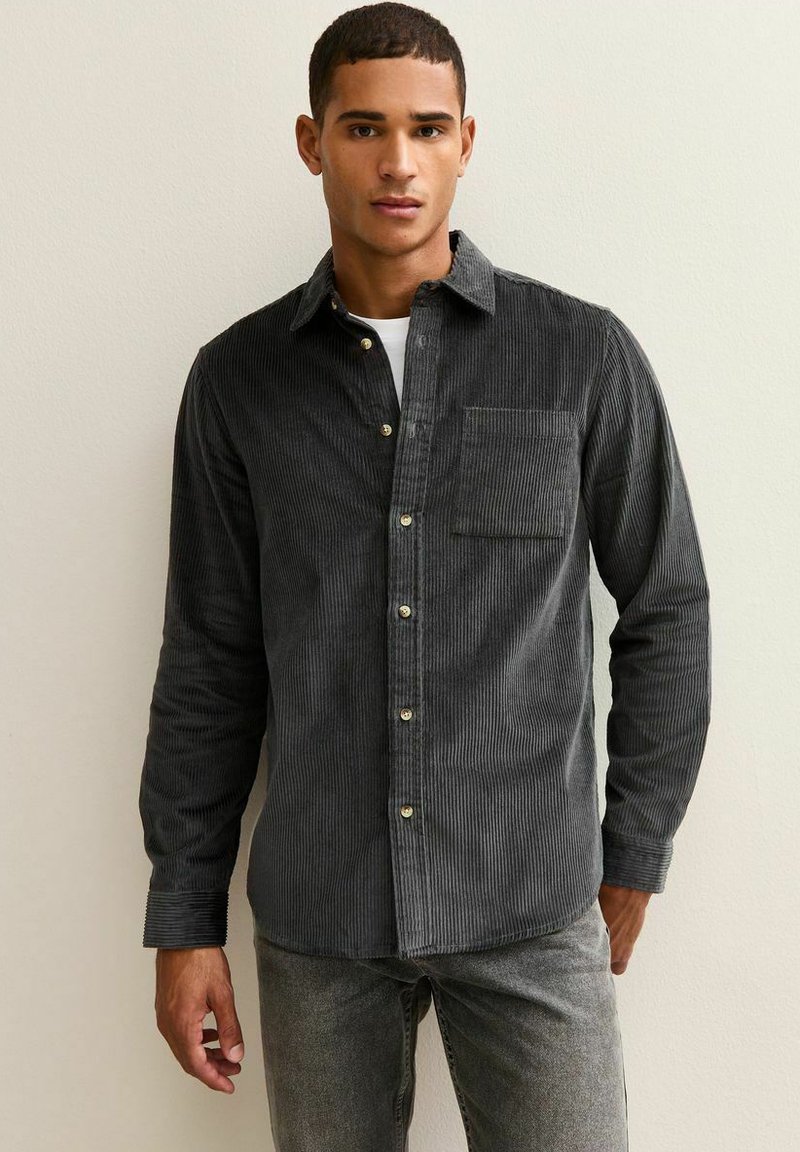 Camisa de corduroy gris oscuro con botones, un único bolsillo en el pecho, rayas verticales prominentes y botones dorados. Mangas largas y corte ajustado.