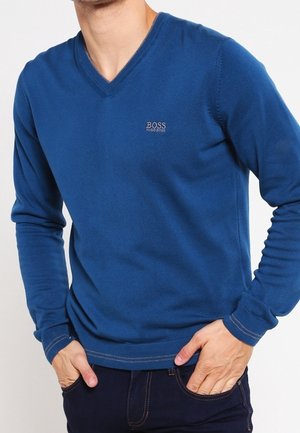 Muž v modrém svetr s výstřihem do V od Hugo Boss s rukama v kapsách tmavě modrých džínů, oříznutý u krku a pasu.