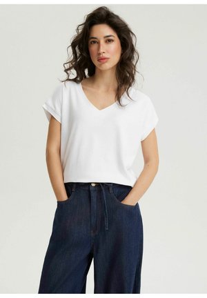 Kobieta o kręconych ciemnych włosach, ubrana w biały krótki top z dekoltem w kształcie litery V oraz ciemnoniebieskie szerokie dżinsy, z rękami w kieszeniach.