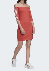 Rode off-shoulder jurk van rekbare stof, met drie witte strepen op elke mouw en een getailleerde pasvorm. G gedragen met witte sneakers.