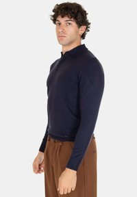 Polo a maniche lunghe blu navy con design colletto, patta con tre bottoni e polsini a costine. Abbinato a pantaloni marroni plissettati.