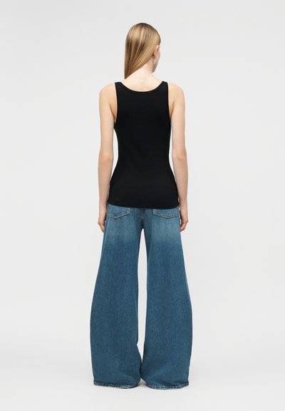 Γυναίκα που στέκεται γυρισμένη με την πλάτη, φοράει εφαρμοστή μαύρη αμάνικη μπλούζα και φαρδιά μπλε τζιν σε μονόχρωμο φόντο.