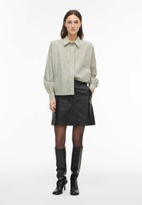 Moteris apsirengusi smėlio spalvos dryžuotą marškinių tipo palaidinę, juodą odinę mini sijoną, permatomas juodas kojines ir juodus aukštakulnius iki kelių.