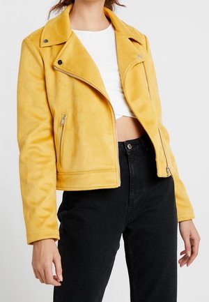 Kobieta w musztardowożółtej zamszowej kurtce motocyklowej na białym krótki topie i czarnych jeansach z wysokim stanem na białym tle.