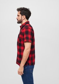 Camisa de franela de manga corta en patrón de cuadros rojo y negro, con cuello abotonado y bolsillo en la parte frontal. Tejido de algodón, ajuste relajado.